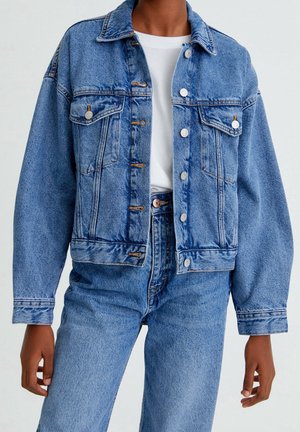 Henkilö, joka pukeutunut siniseen farkkutakkiin valkoisen paidan päällä ja korkeavyötäröisiin sinisiin farkkuihin, seisoo yksiväristä taustaa vasten.