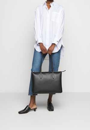 Osoba w białej, dużej koszuli, niebieskich dżinsach, czarnych wsuwanych butach, trzymająca dużą czarną skórzaną torbę typu tote z paskami na ramię.