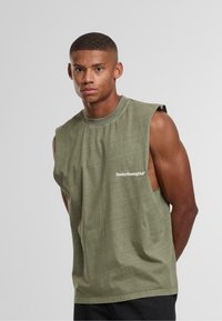 Uomo giovane con capelli corti che indossa una maglietta senza maniche verde oliva con la scritta "Sunday Running Club", in piedi contro uno sfondo chiaro e uniforme.