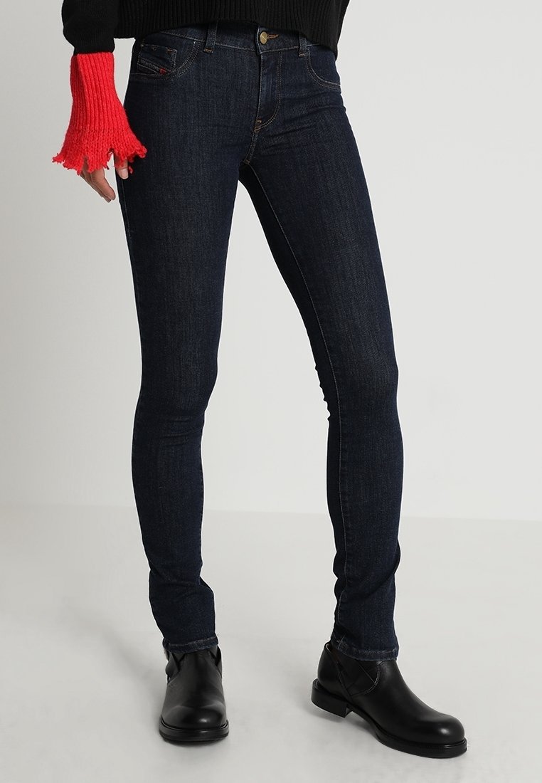 Pantalones vaqueros ajustados de color azul oscuro, fabricados en denim, con un diseño clásico de cinco bolsillos. Se combinan con botas negras hasta el tobillo y un suéter rojo texturizado.