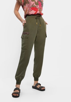 Femme portant un pantalon cargo vert olive avec cordon, poignets élastiques et finitions de poche brodées, assorti à des sandales noires et un haut à fleurs.