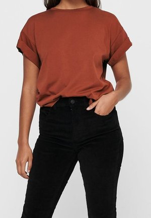 Kvinde iført rustfarvet t-shirt med opkrogede ærmer, stoppet ned i højtaljede sorte bukser med den ene hånd i lommen mod en ensfarvet baggrund.