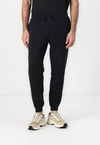 Pantaloni de jogging negri pentru sport, cu talie elastică, detalii cu șnur și picioare conice, asortați cu pantofi de alergare bej.