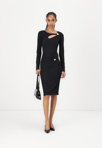 Robe noire à manches longues avec une découpe asymétrique et un nœud latéral, ornée d'un bouton décoratif. Le modèle tient un petit sac à main noir.
