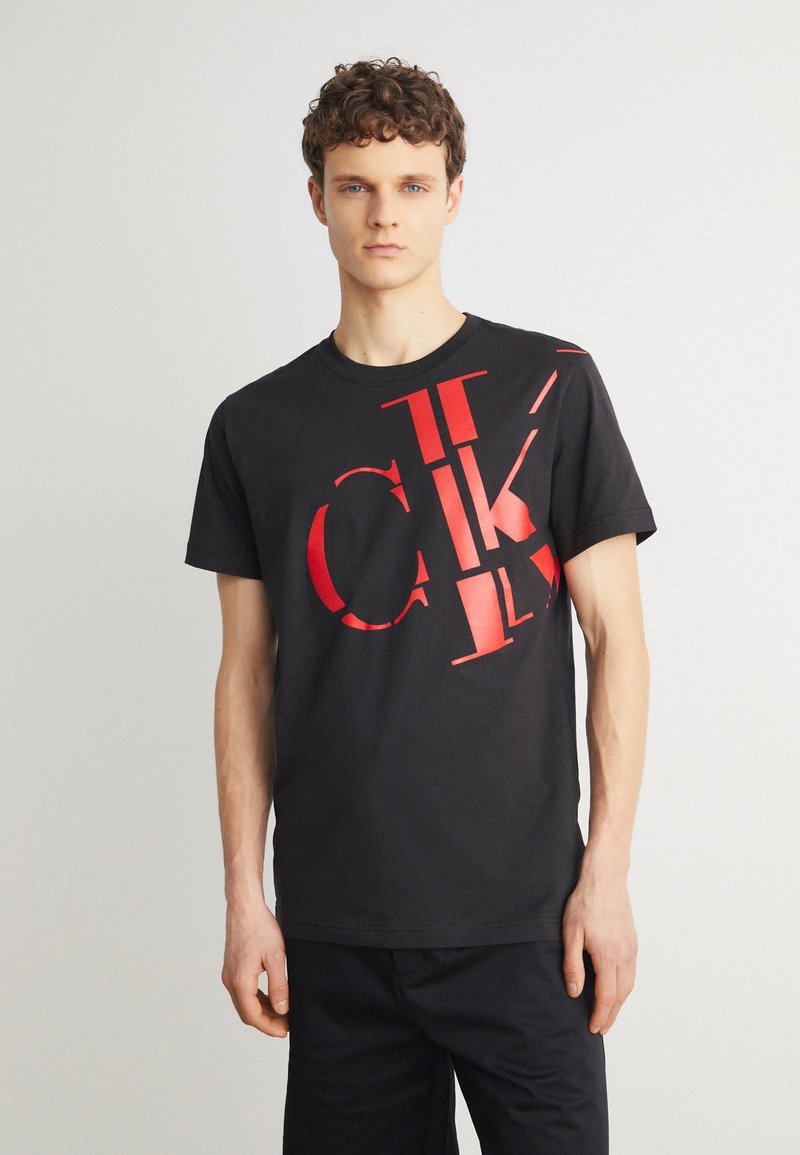 T-shirt en coton noir avec de grandes lettres rouges en block "CK" de tailles variées sur le devant. Design classique à col rond, manches courtes.