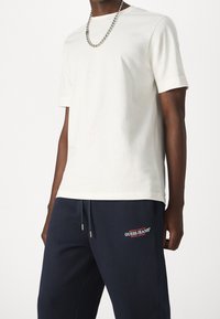 Biała bawełniana koszulka z krótkim rękawem, zestawiona z granatowymi spodniami dresowymi z czerwonym haftowanym logo, metalowymi końcówkami na sznurkówkach.