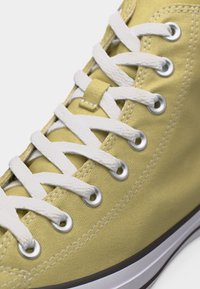 Κοντινό πλάνο ενός κίτρινου καμβά αθλητικού παπουτσιού με λευκά κορδόνια που περνούν μέσα από μεταλλικά πατήματα και λευκή λαστιχένια σόλα με μαύρη ρίγα.