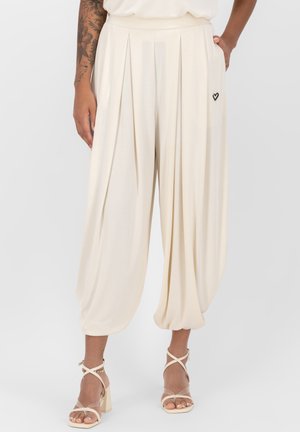 Pantalon plissé large couleur crème avec des ourlets élastiques et un petit détail cœur noir près de la poche, porté avec des sandales à talons blocs à brides.