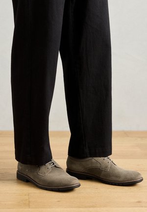 Pantofi din țesătură bej cu un model împletit, vârf rotund și talpă închisă. Asortați cu pantaloni negri tapered, stând pe o podea din lemn.