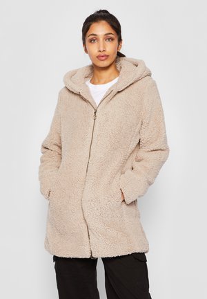 Femeie purtând o geacă fleece bej cu glugă și fermoar, cu mâinile în buzunare, stând în fața unui fundal simplu și deschis.