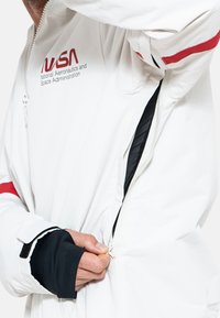 Chaqueta blanca con logo de la NASA, con forro interior negro, detalles en rojo y cierre con cremallera. Los puños tienen agujeros para el pulgar en negro para un mejor ajuste.