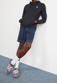 Sudadera negra con capucha y bolsillo frontal, combinada con pantalones cortos de color azul marino. El calzado incluye zapatillas moradas y crema con suelas beige y calcetines blancos.