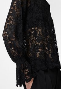 Top preto de renda com padrões florais, corte solto e mangas em forma de sino com punhos franzidos. O tecido é semi-transparente com detalhes intricados.