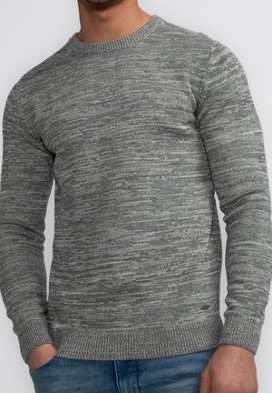 Homme portant un pull en maille gris chiné à manches longues avec des poignets et un bas côtelés, associé à un jean bleu.