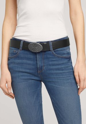 Cinturón negro con una hebilla ovalada metálica texturizada, usado con jeans azules. El cinturón presenta un acabado suave y un diseño de correa ajustable.