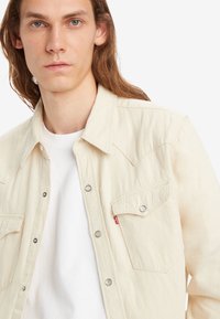 Camisa de denim beige con yugo puntiagudo, dos bolsillos en el pecho, botones a presión grises y una pequeña etiqueta roja en el bolsillo izquierdo. Llevada sobre una camiseta blanca.