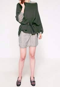 Grön, ribbad oversized topp med ett svart bälte, i kombination med mönstrade shorts i kräm och brunt. Svarta skor med dekorativa detaljer.