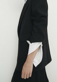 Zwarte blazer met een gestructureerd ontwerp, met witte shirtmanchetten die uit de opgerolde mouwen komen. Zachte stof met nette afwerking.