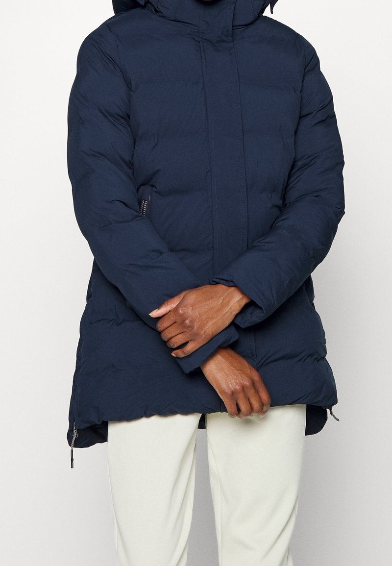 Pikowany granatowy płaszcz z kapturem, zapinany z przodu na zamek, z bocznymi kieszeniami i elastycznymi mankietami, wykonany z gładkiego, ocieplonego materiału.