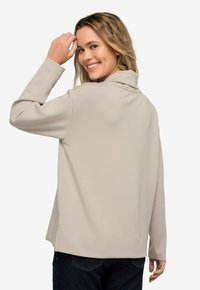 Top de manga larga beige con cuello alto, textura suave y corte relajado; vista posterior mostrando un ligero dobladillo caído y mangas con puños.