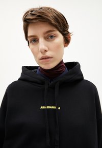 Zwarte hoodie van een katoenen mix met ribgebreide boorden. Heeft een voorkantzak en gele logo-tekst "ARMEDANGELS" gecentreerd op de borst.