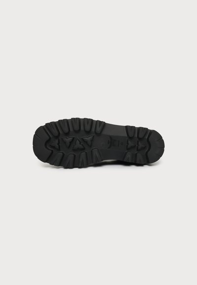 Semelle de botte noire avec des rainures profondes et des motifs texturés sur un fond gris clair.