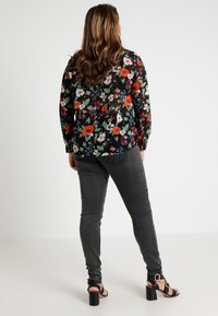Femme aux longs cheveux ondulés portant un blouse à fleurs noire, un jean skinny gris foncé et des sandales à talons noires, se tenant de dos par rapport à l'appareil photo.