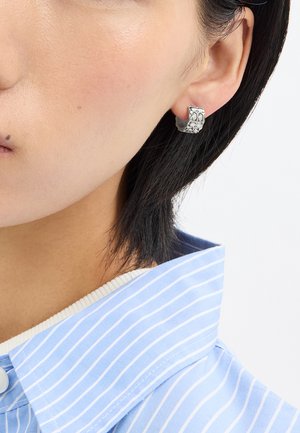 Boucles d'oreilles en argent avec une finition texturée, présentant un design géométrique. La personne qui les porte a les cheveux noirs raides et est vêtue d'une chemise à rayures bleues et blanches.