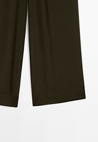Pantalons larges en olive foncé avec un ourlet propre sur fond blanc uni, montrant la partie inférieure de la jambe.