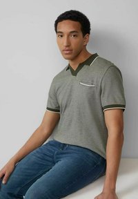 Graues Poloshirt mit kurzen Ärmeln und einem dunkleren grünen Kragen, weißen Akzenten und einer kleinen Brusttasche. Getragen mit blauen Jeans.