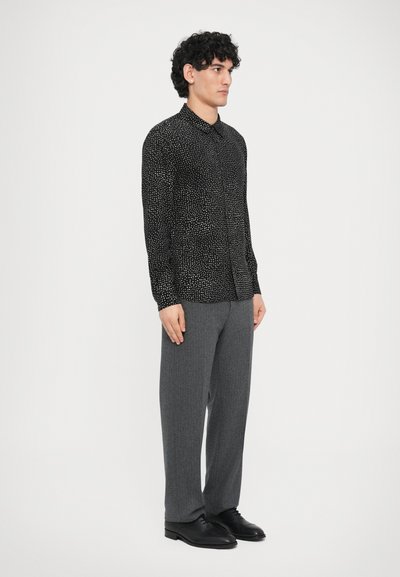 Schwarzes Langarmshirt mit kleinen weißen Punkten, Hemdkragen mit Knöpfen, kombiniert mit grauer maßgeschneiderter Hose und schwarzen eleganten Schuhen.