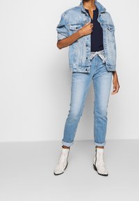 Giacca di jeans di un azzurro chiaro, indossata sopra una maglietta navy, abbinata a jeans azzurri chiari e stivaletti alla caviglia in bianco con motivo serpente e cerniere.