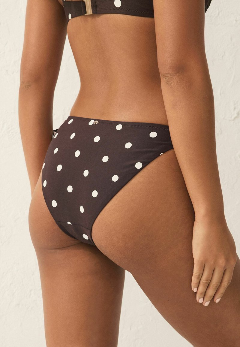 Costum de baie de culoare neagră, cu buline albe. Are un croi jos și o textură netedă, accentuată de un mic detaliu metalic pe lateral.