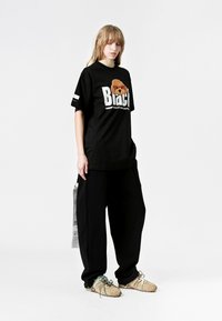 Camiseta negra de gran tamaño con un gráfico de un oso y texto en blanco, combinada con pantalones holgados negros y zapatillas beige con cordones verdes y marrones.