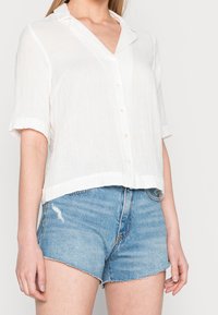 Camicia bianca a maniche corte con bottoni, realizzata in tessuto leggero, che presenta un colletto e una vestibilità rilassata, abbinata a pantaloni corti in denim sfilacciati.