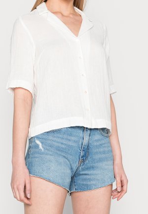 Camicia bianca a maniche corte con bottoni, realizzata in tessuto leggero, che presenta un colletto e una vestibilità rilassata, abbinata a pantaloni corti in denim sfilacciati.