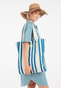 Gestreepte tote bag met blauw en witte strepen, gemaakt van stof met platte handvatten. De draagster is gekleed in een bijpassende gestreepte shirt en rok.