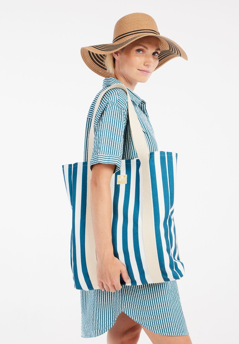 Gestreepte tote bag met blauw en witte strepen, gemaakt van stof met platte handvatten. De draagster is gekleed in een bijpassende gestreepte shirt en rok.