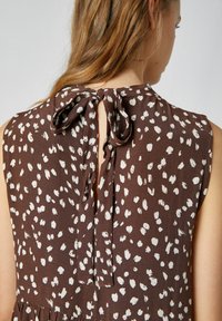 Brun ärmlös topp med ett vitt prickigt mönster, med ett knyt bak i nackpartiet och ett lätt texturerat tyg.
