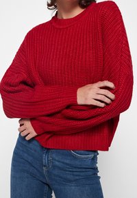 Donna che indossa un maglione rosso a maglia grossa con maniche lunghe e jeans in denim blu, con le braccia incrociate in vita.