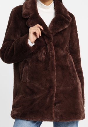 Nainen, joka käyttää tummanruskeaa pehmeää tekoturkistakkia valkoisen pooloneuleen ja sinisten farkkujen päällä.