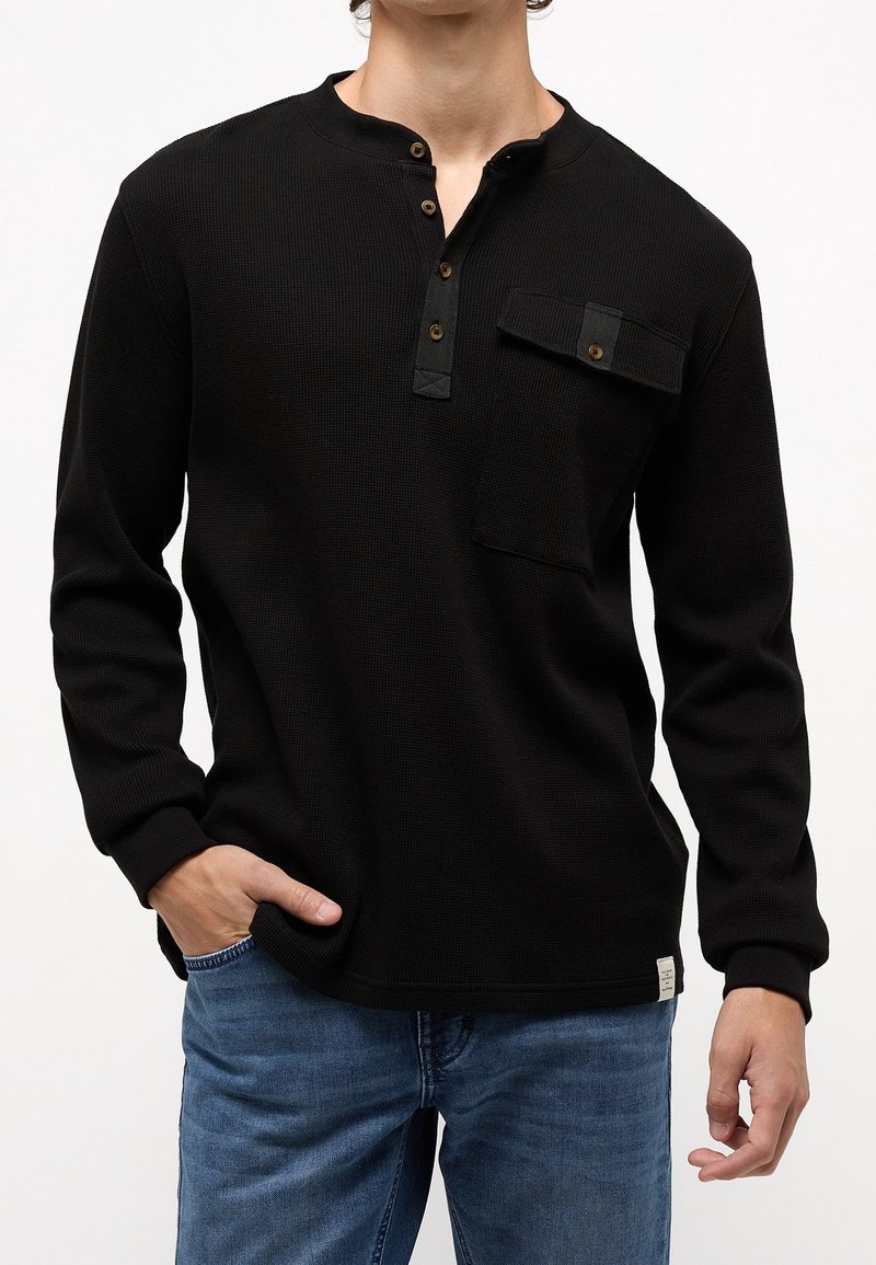 T-shirt henley à manches longues noir en tissu tricot texturé, avec un col à boutons partiel, une poche sur la poitrine et une étiquette sur le côté.