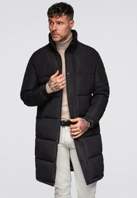 Sort polstret jakke med høj krave, lange ærmer og trykknaplukning, båret over en beige turtleneck. Har en kviltet tekstur.