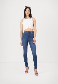 Mulher em pé, vestindo uma parte de cima branca sem mangas, jeans skinny azuis de cintura alta, saltos roxos com tiras e óculos de sol na cabeça.