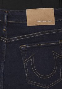 Dark denim jeans with a tan leather patch labeled "TRUE RELIGION" and contrasting gold stitching on pockets and seams.