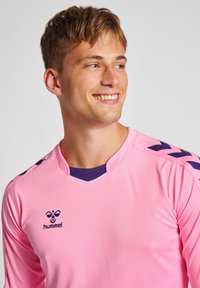 Jersey sportivo a maniche lunghe rosa con colletto e logo viola. Presenta accenti a chevron neri sulle spalle. Materiale morbido e leggero.