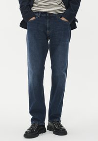 Ravne temno modre jeans hlače, ki jih nosi oseba z rokama v žepih, v kombinaciji s črnimi vezalkami in prugastim srajcem ter mornarsko modrim jopičem.