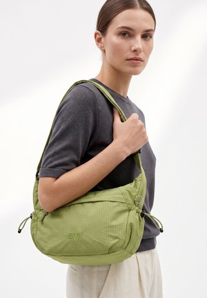 Donna con top grigio a maniche corte che porta una borsa a tracolla verde chiaro con il logo "envy" sul davanti, su uno sfondo semplice.