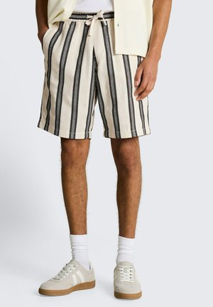 Mężczyzna w beżowo-czarnych szortach w pionowe paski z troczkiem, białych skarpetkach, kremowych sneakersach i kremowym swetrze z kieszeniami.