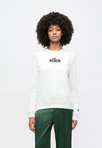 Γυναίκα με σγουρά μαλλιά που φοράει λευκό φούτερ Nike και πράσινο παντελόνι, στέκεται μπροστά σε απλό λευκό φόντο.